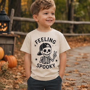 Puede incluir: Una camiseta beige de manga corta con un gráfico negro de un esqueleto que lleva una gorra de béisbol y hace el signo de la paz. El texto "FEELING SPOOKY" está impreso en negro encima y debajo, decorado con pequeñas estrellas.