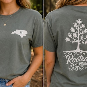 K&ouml;nnte beinhalten: Ein schiefergr&uuml;nes Kurzarm-T-Shirt mit einer wei&szlig;en Grafik des Umrisses von North Carolina und den Buchstaben NC auf der Brust sowie einer gro&szlig;en wei&szlig;en Kieferngrafik mit Wurzeln und dem Text Rooted IN THE PINES auf der R&uuml;ckseite.