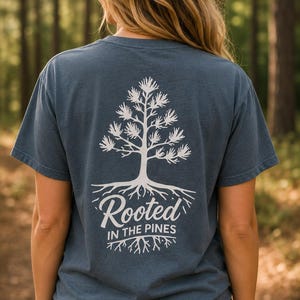K&ouml;nnte beinhalten: Ein schieferblaues Kurzarm-T-Shirt mit einer wei&szlig;en Grafik auf der R&uuml;ckseite. Das Design zeigt eine Kiefer mit sichtbaren Wurzeln und den Text "Rooted IN THE PINES", der in wei&szlig;er Schrift unter der Baumillustration gedruckt ist.