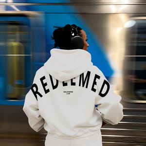 May include: White hoodie with the word "REDEEMED" printed in large black letters across the back. The text "GALATIANS 2:20" is printed below the word "REDEEMED". The person is wearing black headphones.