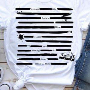 Può includere: T-shirt bianca con un design grafico nero. Il design presenta il testo "release the Epstein files, because we have nothing to hide" e "Donald J. Trump". È incluso anche un timbro "CLASSIFIED".