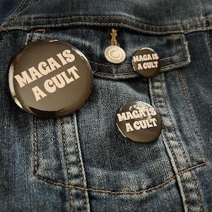 Puede incluir: Un conjunto de botones negros con el texto blanco "MAGA IS A CULT" sobre una chaqueta vaquera. Los botones varían en tamaño, con uno grande y varios más pequeños. La chaqueta tiene un botón y un bolsillo.