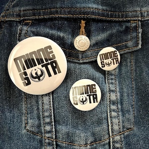May include: Three white round buttons with the word "MINNESOTA" in black block letters and a black graphic of a bird. The buttons are on a blue denim jacket.