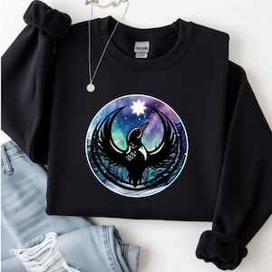 May include: Black sweatshirt featuring a circular graphic of a bird with outstretched wings, a star, and a night sky background. The bird has a red eye and is surrounded by a forest silhouette. The sweatshirt has long sleeves and a crew neck.