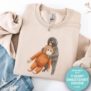 May include: A beige sweatshirt with a graphic of two monkeys hugging. One monkey is grey, and the other is orange. The sweatshirt has a crew neck and long sleeves. The image also includes a button that says "AVAILABLE AS A T-SHIRT SWEATSHIRT HOODIE & MORE."