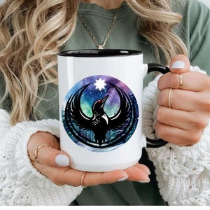 May include: A white ceramic mug with a black handle and interior rim. The front features a circular graphic depicting a black bird with red eyes, wings spread wide, set against a vibrant purple and blue nebula background with a bright white star.