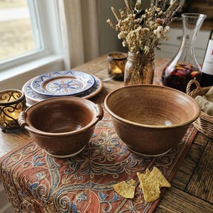 May include: Two handmade ceramic bowls with a smooth, earthy brown glaze. The smaller bowl features two side handles, while the larger bowl has a wide, open rim. Both bowls have a clean, white circular base and a simple, functional design.