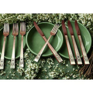May include: A set of vintage silverware, including forks and knives, arranged on green plates. The silverware has a decorative design on the handles and a copper-colored finish. The scene is set on a bed of greenery and white flowers.