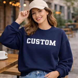 Young woman wearing navy blue sweatshirt with white varsity CUSTOM text design, lifestyle mockup in urban outdoor setting showing personalized sweatshirt styling, Nicheville custom