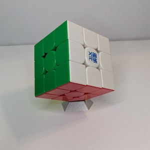 May include: A three-dimensional puzzle cube with green, white, and red sides. The cube is resting on a small, silver-colored stand. The cube has a logo on one of its white squares.