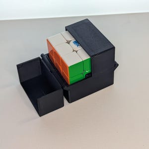 May include: A black, hinged case with a partially visible, multi-colored Rubik's Cube inside. The cube has white, orange, green, and blue sides. The case is open, revealing the cube and its protective storage.