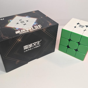 May include: A black box with gold accents and the word "MOYU" on top, next to a solved green, white, and red cube puzzle. The box has Chinese text and a graphic of a cube. The cube has Chinese characters on each side.
