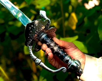 Puede incluir: Una espada con una empuñadura de metal plateado pulido y un mango negro con un patrón en espiral de pequeños tachones plateados. La guarda es ancha y ornamentada con un arco curvo, y la hoja es recta, metálica y reflectante.
