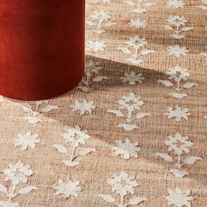 May include: A tan jute rug with a repeating white floral pattern. A cylindrical, rust-colored velvet ottoman is in the foreground, with a white-topped table and terracotta vase visible. The rug has a natural, textured appearance.