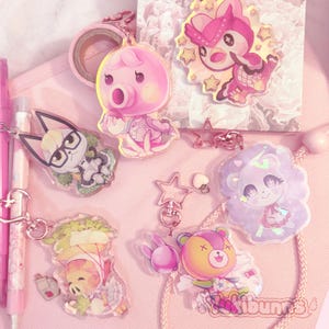 Op de afbeelding: Een verzameling roze en transparante acryl bedels met cartoon dierenfiguren. De bedels bevatten een roze octopus, een hert, een kat, een beer, een varken en een eenhoorn. Andere items zijn een rol roze washi tape, pennen en stervormige bedels.