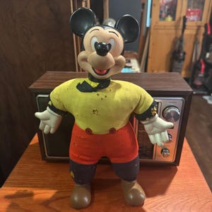 May include: Vintage Mickey Mouse doll with a black head and ears, a yellow shirt, red shorts, and white gloves. The doll is standing in front of a brown radio on a wooden surface. The doll has a black bow tie and brown shoes.