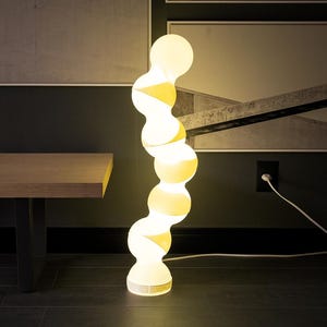 Peut inclure: Une lampe de sol blanche moderne avec un design incurvé unique. La lampe est composée d'une série de luminaires sphériques interconnectés qui émettent une lueur jaune chaude. La lampe est branchée sur une prise murale. La lampe mesure environ 152 cm de haut.