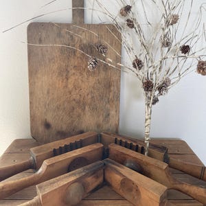 May include: A collection of antique wooden kitchen tools, including several butter molds and a large cutting board. The tools are arranged on a wooden surface, with a decorative branch with pinecones in the background. The wood has a warm, aged appearance.