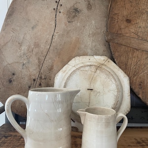 May include: Two off-white ceramic pitchers with a glossy finish and subtle horizontal ridges. The larger pitcher is on the left, and the smaller one is on the right. Both feature a single handle and a pouring spout, showing fine surface crazing.