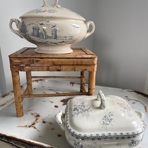 May include: Two antique cream-colored serving dishes with lids. One is round with a landscape scene, the other is square with floral patterns. Both have handles and are on a bamboo table and a white tray.