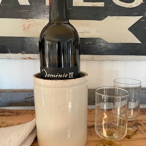 May include: A wine bottle with a black label that reads "Dominio IV" is placed in a cream-colored ceramic wine chiller. Two clear glasses with amber-colored square bases sit on a wooden surface. A white cloth is on the left.