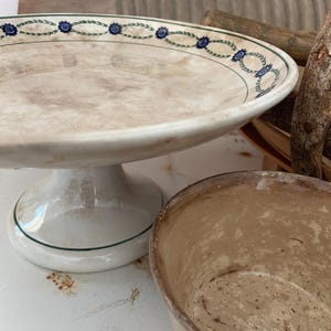 May include: A ceramic pedestal bowl with a wide, shallow basin featuring a cream-colored surface and a decorative rim pattern of blue flowers and green leaves. Beside it sits a smaller, weathered, light brown bowl with a textured interior finish.