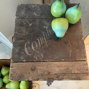 May include: A rustic wooden crate with the word "Compto" stenciled on the top. Three green pears rest on the crate's surface. A basket filled with green pears is visible below.