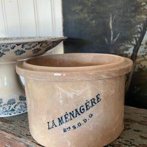 May include: A cylindrical beige stoneware crock with a wide rim. The exterior features dark blue printed text that reads LA MÉNAGÈRE and Bté S.G.D.G. The surface has a smooth, glazed finish with subtle marbled color variations throughout the clay body.