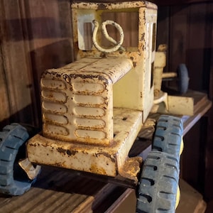 May include: Vintage toy tractor in yellow and blue, showing signs of wear and rust. The tractor has a detailed front grill, a steering wheel, and blue tires. The toy is made of metal and sits on a wooden shelf.