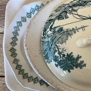 May include: A stack of antique white ceramic serving dishes. The top dish has a lid with a teal landscape design. The other dishes have teal geometric patterns around the edges. The dishes have a rustic, vintage aesthetic.