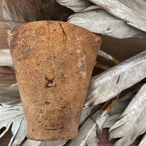 May include: A weathered, cone-shaped, brown pottery piece with visible cracks and a stamped emblem. The emblem reads "BATTERSEA ENGLAND". The piece has a rustic, aged appearance, suggesting it is an antique or vintage item.