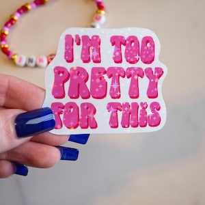 May include: A pink and white sticker with the text "I'M TOO PRETTY FOR THIS" in bubble letters. The sticker has a glittery effect. A beaded bracelet with pink, white, and gold beads is in the background.