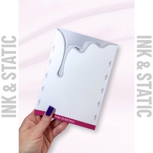 May include: A white notepad with a silver liquid design dripping from the top. Small lightning bolt graphics are scattered across the page. The bottom edge has a magenta stripe with the text "STAY ELECTRIC." The words "INK & STATIC" are printed vertically on the left and right sides.