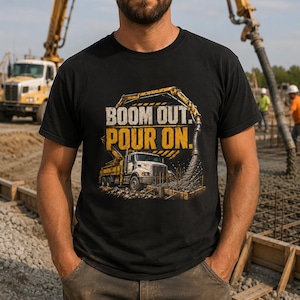 May include: A black short-sleeved t-shirt featuring a graphic of a yellow concrete pump truck pouring cement. The text "BOOM OUT. POUR ON." is printed in bold yellow and white letters above the truck illustration on the front of the garment.