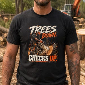 May include: A black t-shirt featuring a graphic illustration of a logger using a chainsaw on a tree trunk. The text "TREES DOWN" appears in white above the logger, and "CHECKS UP" is printed in white and orange below the graphic design.