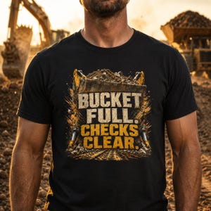 May include: A black short-sleeved t-shirt featuring a graphic of an excavator bucket filled with dirt. The text BUCKET FULL. CHECKS CLEAR. is printed in a distressed yellow and white font over the image of the heavy machinery bucket.
