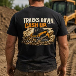 May include: A black short-sleeved t-shirt featuring a graphic on the back of a yellow skid-steer loader moving dirt. The text above the illustration reads "TRACKS DOWN. CASH UP." in white and orange lettering against a dusty background.