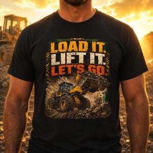 May include: A black short-sleeve t-shirt featuring a graphic of a yellow construction loader moving dirt. The text "LOAD IT. LIFT IT. LET'S GO." is printed in bold yellow, white, and orange letters above the heavy machinery illustration.