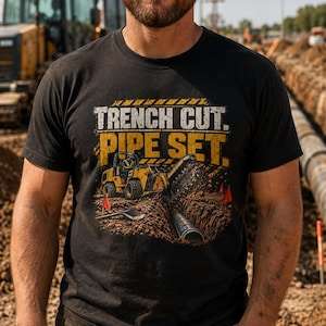 Op de afbeelding: Een zwart t-shirt met een grafische print van een gele sleuvengraver die in de aarde graaft. De tekst TRENCH CUT. PIPE SET. staat in grote gele en witte letters boven de bouwmachine en een buisgedeelte in de grond gedrukt.
