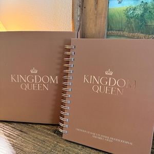 May include: Two brown journals with gold spiral binding and the words "KINGDOM QUEEN" printed on the cover. One journal is closed, and the other is open with the text "3 MONTHS OF DAILY PLANNER, PRAYER JOURNAL AND BIBLE STUDY."
