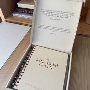 May include: A white spiral-bound planner with the words "KINGDOM QUEEN" in rose gold lettering, inside a white box. The box lid has text from Ephesians 5:15-17. The planner is labeled "3 MONTHS OF DAILY PLANNER, PRAYER JOURNAL, AND BIBLE STUDY."