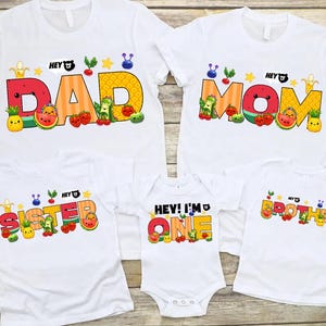 May include: White t-shirts with colorful fruit-themed graphics. The shirts say "DAD," "MOM," "SISTER," "BROTHER," and "HEY! I'M ONE." Each shirt features a different fruit design, including watermelon, pineapple, and strawberries.
