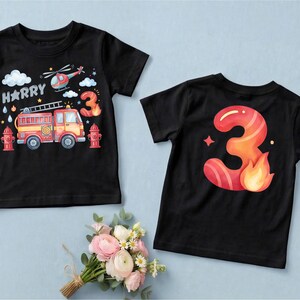 May include: Two black t-shirts. One features a colorful illustration of a fire truck, helicopter, and the name "Harry" with a number "3". The other shirt has a large number "3" with flames. Both shirts are for a child's birthday.