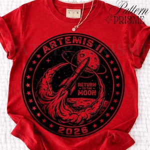 May include: Red t-shirt with a black and red graphic. The design shows a rocket heading towards a planet, with the text "ARTEMIS II" and "RETURN TO THE MOON 2026". A gold necklace is visible.