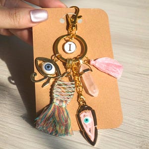 May include: Gold-tone keychain with an initial "I" charm, an eye of Horus charm, a multi-colored tassel, a pink crystal, and a pink and black evil eye charm. A pink tassel and a gold clasp complete the design. The keychain is attached to a brown card.