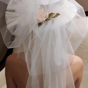 May include: A white bridal hat with a wide brim, adorned with a large tulle bow and a delicate blush-colored rose. A sheer veil cascades down, creating a soft, romantic look. The hat is a classic accessory for a wedding.