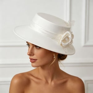 May include: A white hat with a wide brim and a cylindrical crown. The hat has a satin ribbon band and is decorated with a fabric rose and delicate embellishments. This elegant hat is suitable for special occasions.