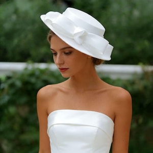 Peut inclure: Un chapeau blanc avec un large bord et un grand nœud. Le chapeau est fait d'une matière lisse, peut-être satinée. Le chapeau est de style classique, adapté aux événements formels ou aux mariages.