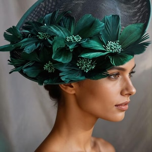Peut inclure: Un chapeau vert foncé et noir à large bord, orné de grandes fleurs en plumes vert émeraude et de petits détails perlés. Le chapeau a un dessus en résille, idéal pour les événements formels.