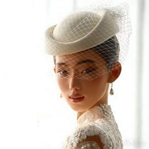 Op de afbeelding: Een crèmekleurige pillbox hoed met een bijpassende sluier. De hoed heeft een afgeronde kroon en een licht opstaande rand. De sluier is een fijn gaas dat over het gezicht drapeert. De hoed maakt deel uit van een bruids- of formele outfit.